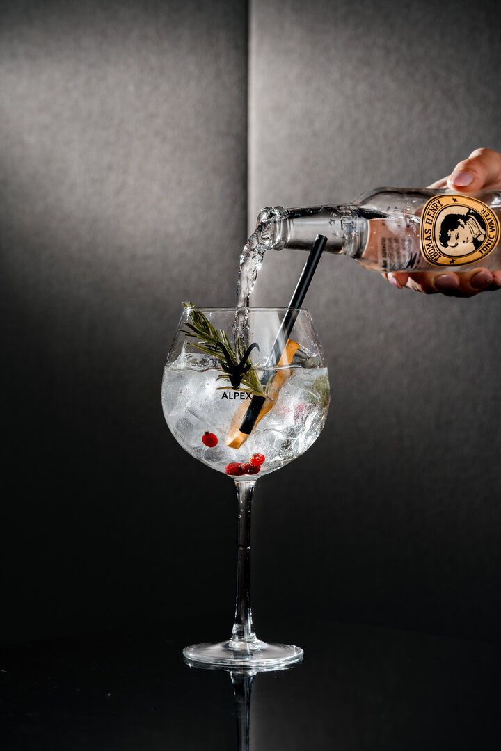 A drink being poured into a glass with ice, garnish, and berries, against a dark background.