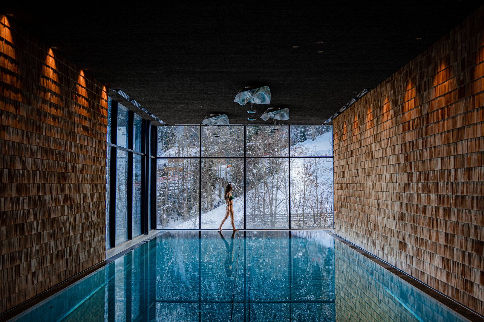 A woman in a swimsuit walks beside an indoor pool with wooden walls, large floor-to-ceiling windows, and a snowy winter landscape visible outside.