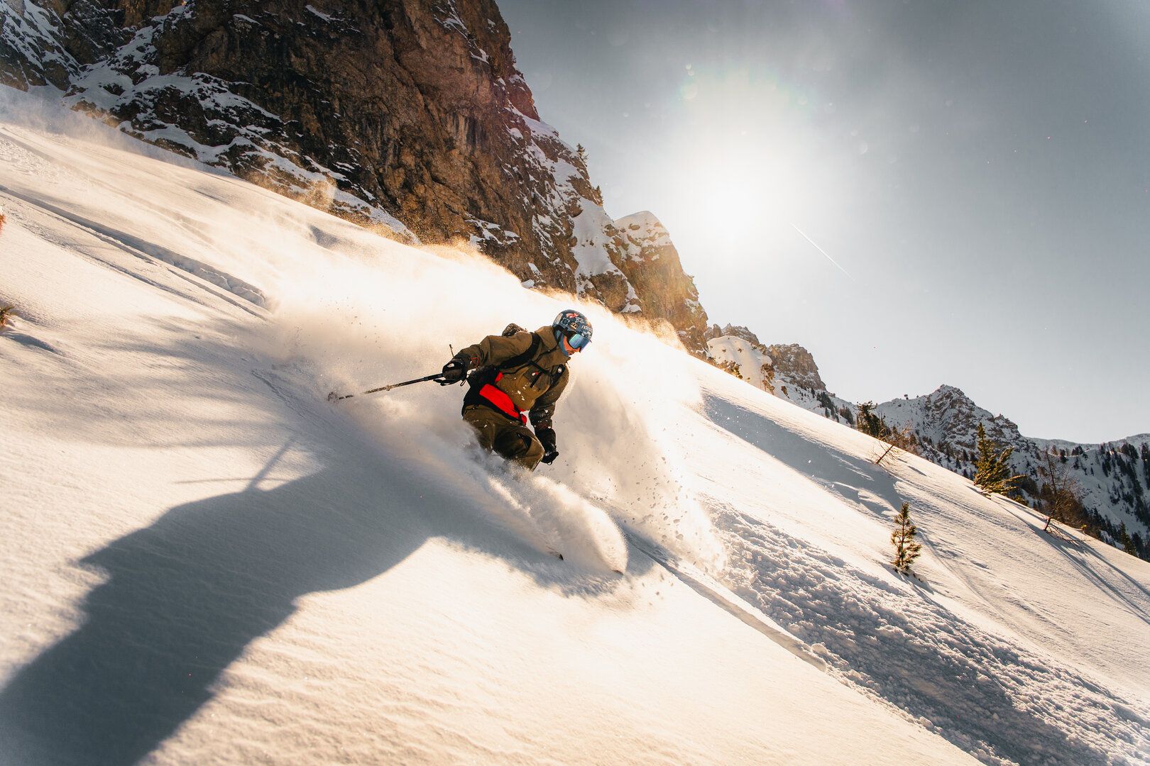 Skier making a turn on a snowy mountain with sunlight and mountains in the background.